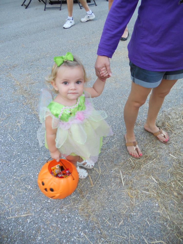 Young girl dressed in bright green and pink dress holding woman's hand and orange jack-o-lantern bucket
